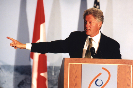 Bill Clinton speaking at podium in front of Canada and US flags